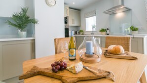 A modern kitchen with a wooden table set for a meal, featuring a bottle of Brancott Estate wine, a candle, grapes, and cheese. - Bryntirion Wyn - sleeps 4 guests  in 2 bedrooms (Aberffraw)