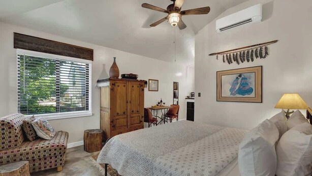Un dormitorio con una cama, un armario de madera, una mesa de comedor con sillas, un ventilador de techo y una ventana con persianas.