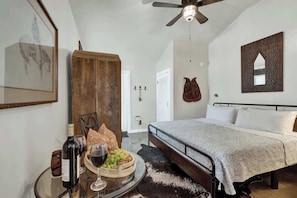 A bedroom with a bed, a ceiling fan, a wooden wardrobe, a framed artwork, and a table with wine and fruit. - Casa Salvia Tejas Suite Walking Distance to MainSt! (Fredericksburg)