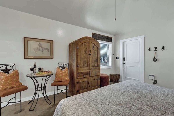Un dormitorio con una cama, un armario de madera, una mesa redonda con sillas, un cuadro enmarcado en la pared y una puerta blanca.
