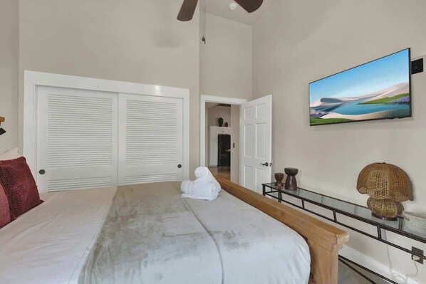 A bedroom with a large bed, white closets, a flat-screen TV, and a view of a landscape painting.