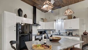 A modern kitchen with white cabinets, a stainless steel refrigerator, a wooden island with a fruit platter and wine glasses, and pendant lighting.
