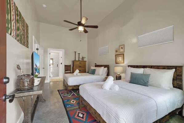 A hotel room with two beds, a ceiling fan, a wooden headboard, a small table with a decorative item, and a colorful rug.