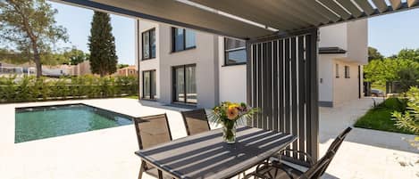 A modern outdoor patio with a pool, a table with chairs, and a pergola overhead.