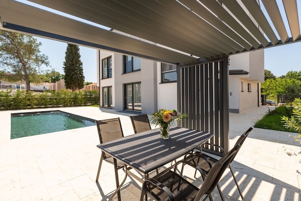 A modern outdoor patio with a pool, a table with chairs, and a pergola overhead.