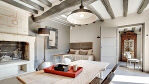 A rustic room with exposed wooden beams, a fireplace, a bed with a wooden headboard, a dining table with a red tray, and a large mirror. - Les Suites Du Cha Teau (Candes-Saint-Martin)