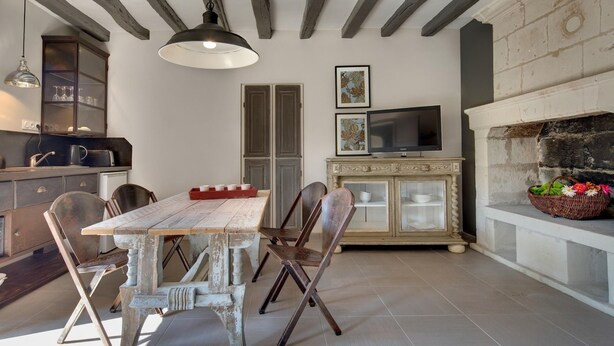 A dining area with a rustic wooden table and chairs, a television on a cabinet, and a fireplace with a basket of flowers. - Les Suites Du Cha Teau (Candes-Saint-Martin)