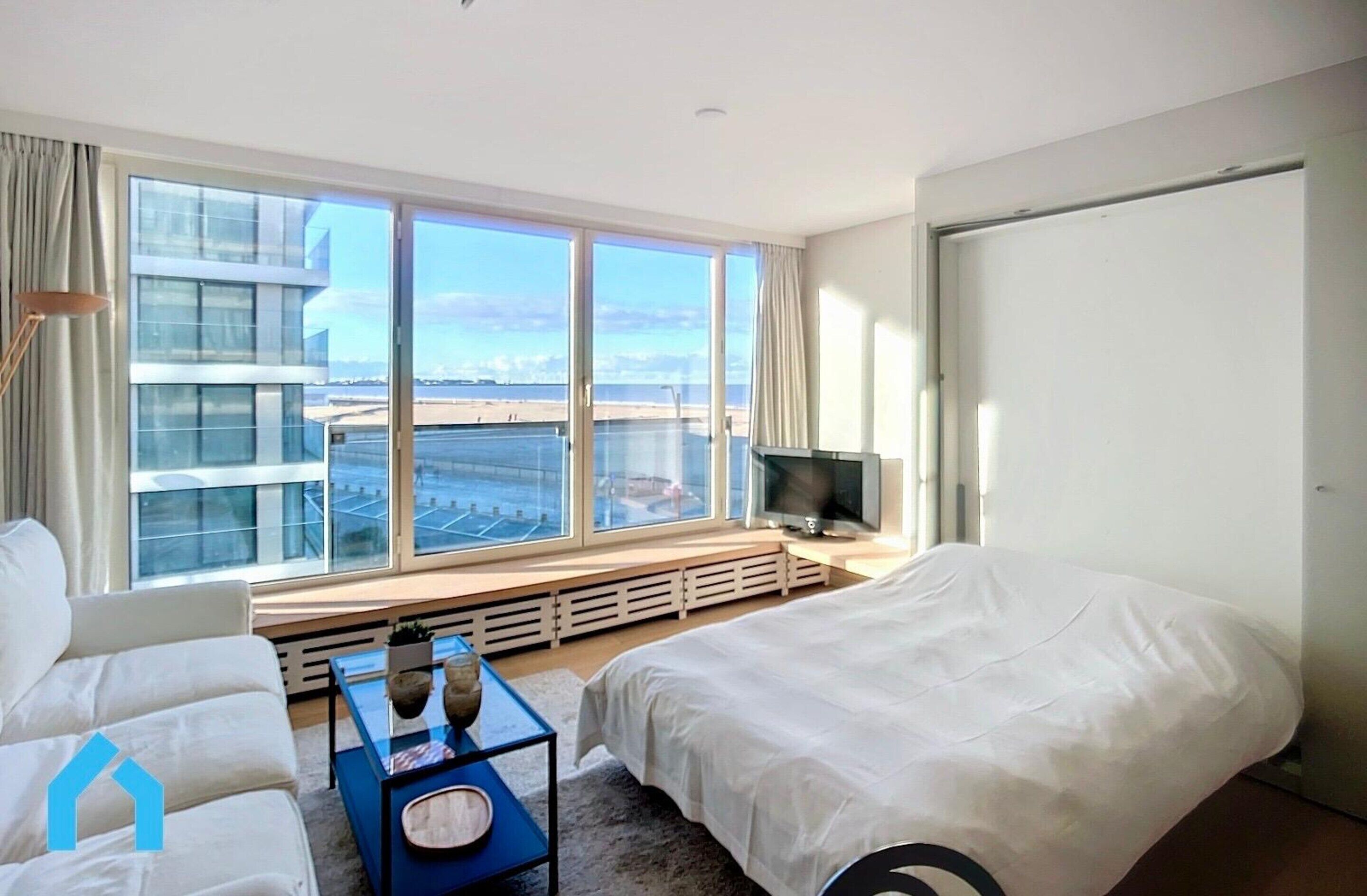 Ein modernes Hotelzimmer mit einem großen Fenster, das Blick auf einen Strand und Gebäude bietet, ein Bett, eine Sitzecke und ein kleiner Tisch mit einem Fernseher.