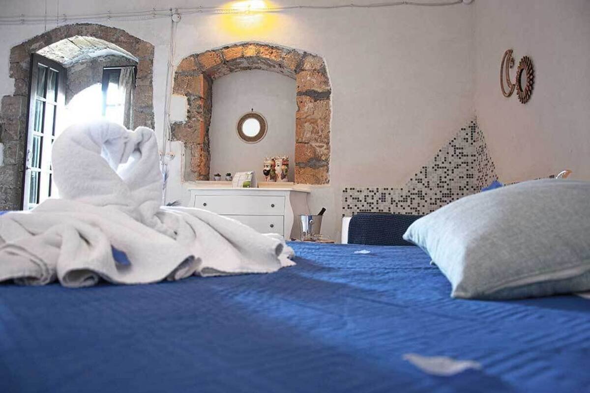 A bedroom with a blue bedspread, a white blanket, a stone archway, a round mirror, and a wall with a mosaic pattern.