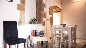 A room with a black chair, a small table with a white tablecloth, and a white dining table with chairs. There is a brick wall with exposed stone and a window above the table. - Il Casale Rooms&Apartment (Noto)