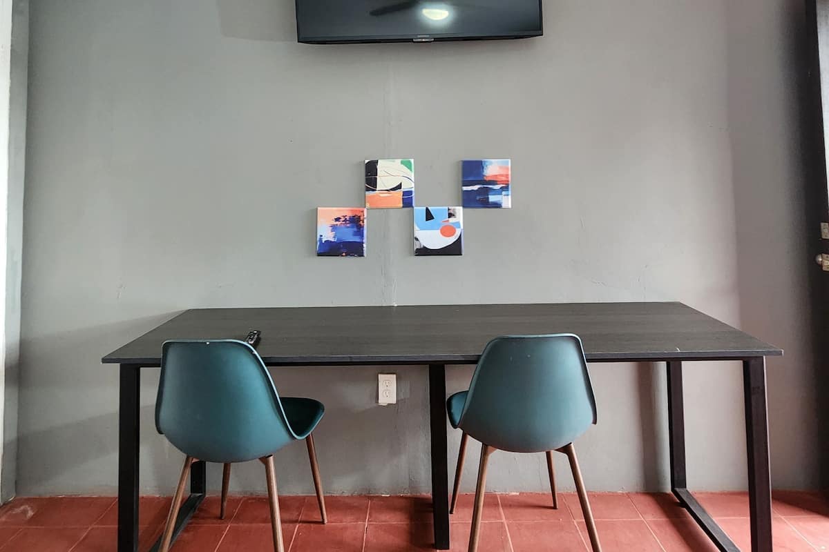 Una stanza con una parete grigia, una TV a schermo piatto montata sopra un tavolo con due sedie color petrolio e tre piccoli quadri incorniciati sotto la TV.