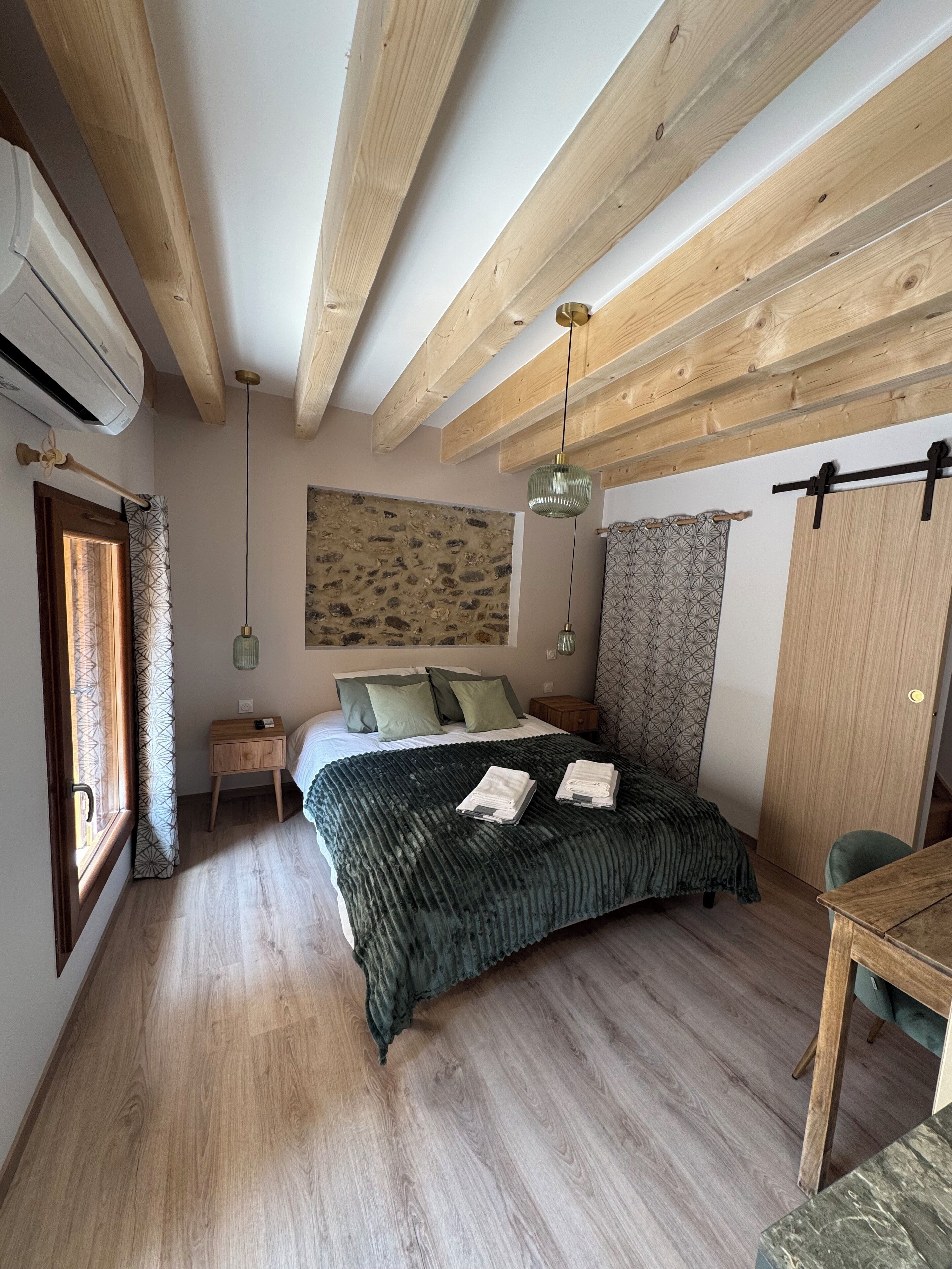 Un dormitorio con techo de madera, una cama con manta verde, una mesita de noche y una lámpara colgante, además de una ventana con cortinas.