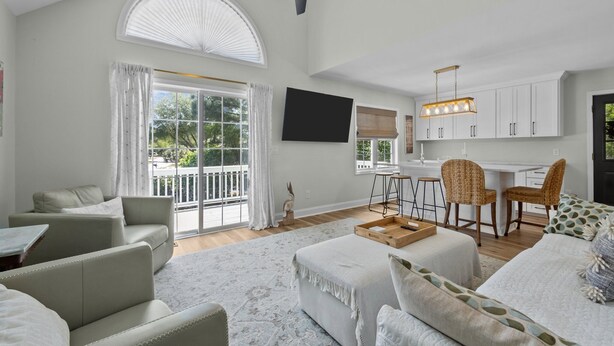 A spacious living room with a large window, a ceiling fan, and a kitchen area with white cabinets and bar stools. - 3BR - Dogs OK! Near the Pointe ~ Hello Sunshine (Emerald Isle)