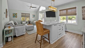 A spacious living room with a large white sofa, a wicker chair, and a dining area with a white island and bar stools. - 3BR - Dogs OK! Near the Pointe ~ Hello Sunshine (Emerald Isle)