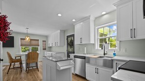 A modern kitchen with white cabinetry, a central island, and a dining area with wicker chairs. - 3BR - Dogs OK! Near the Pointe ~ Hello Sunshine (Emerald Isle)