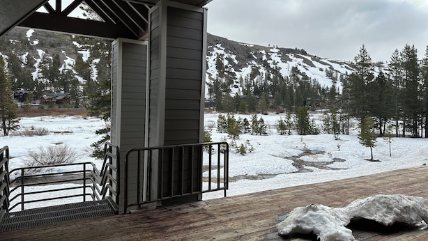 A wooden deck with a metal railing, overlooking a snowy landscape with trees and a mountain in the distance.