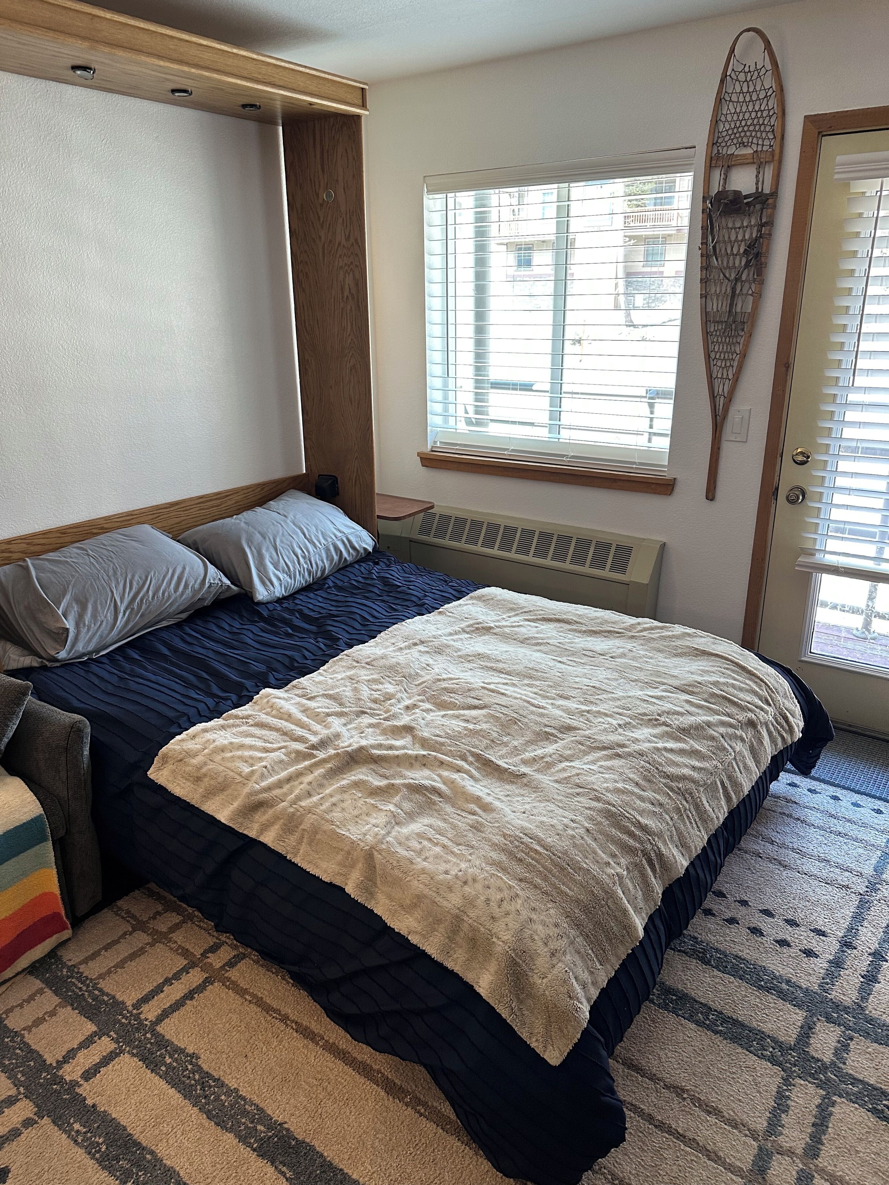 A bedroom with a bed, a window with blinds, a wall-mounted shelf, and a door with a decorative item.