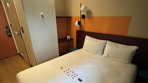 A hotel room with a large bed, a bedside table, a wall-mounted lamp, and a door with a key card lock. - Unisotel (Bron)