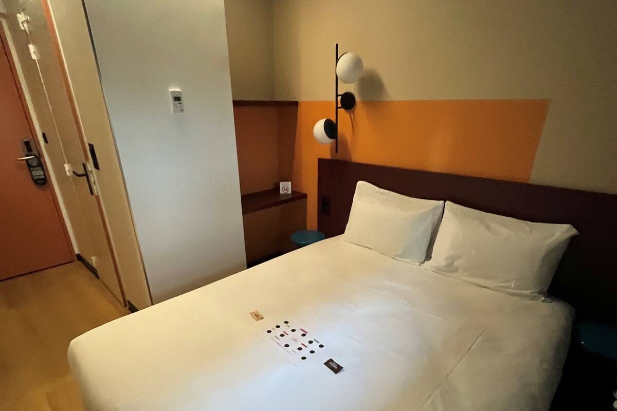 A hotel room with a large bed, a bedside table, a wall-mounted lamp, and a door with a key card lock.