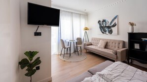 Egy modern nappali, melyben található egy síkképernyős televízió, egy kanapé, egy étkezősarok székekkel és egy nagy növény.