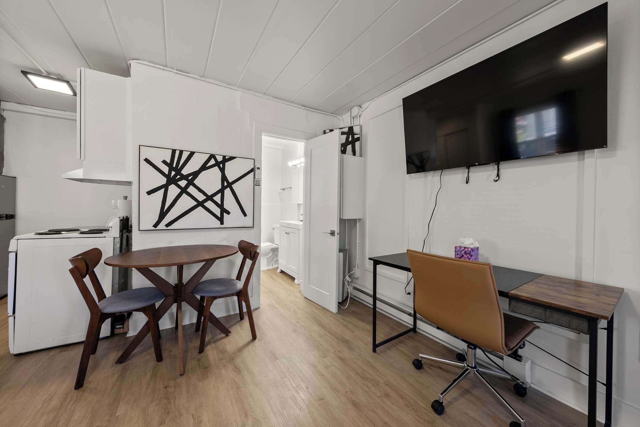 Una cocina moderna con armarios blancos, una mesa de comedor con sillas, un televisor de pantalla plana en la pared y un pequeño espacio de trabajo con escritorio y silla.