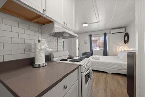 A compact kitchen with a stove, oven, and white cabinets. There is a bed with white bedding in the adjacent room.
