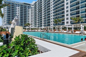 A hotel pool area with lounge chairs and umbrellas, surrounded by a large building with multiple balconies.