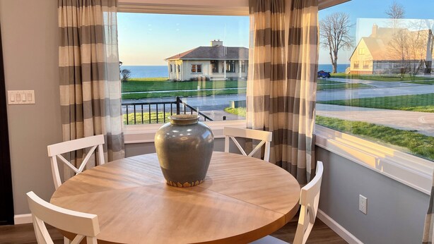 Un coin repas avec une table ronde, des chaises blanches, un grand vase et une vue sur une maison et une rue, le tout visible à travers des rideaux.