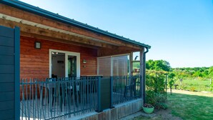 A wooden cabin with a covered porch, a fenced patio, and a view of a lush green landscape.