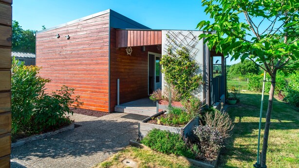 A modern building with a wooden exterior, a small entrance, and a garden area with various plants and a tree.