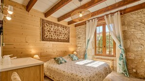 A cozy bedroom with a wooden ceiling, a stone wall, a large bed with floral bedding, a bedside table with a lamp, and a window with curtains. - Le Four - Piscine Privée (Thénac)