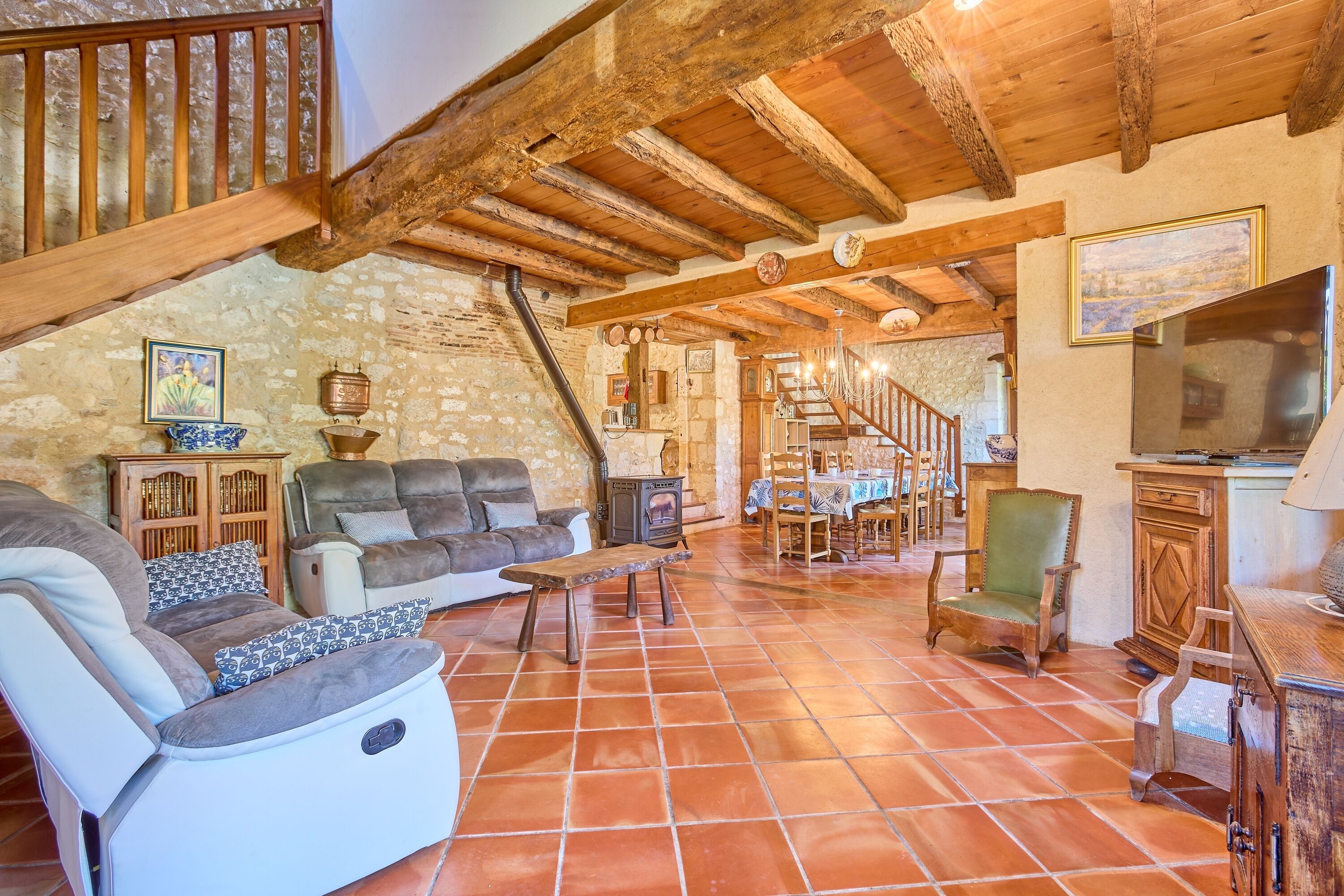 A rustic interior with a stone wall, wooden beams, a sofa, a dining table, and a television.