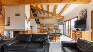 A spacious living room with a black leather sofa, wooden beams, a fireplace, and a large window overlooking a mountain view.