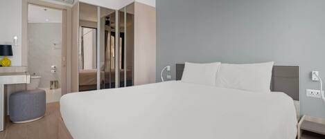 A modern hotel room with a large bed, a bedside table, a small stool, a mirror, and a walk-in closet.