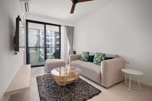 A modern living room with a sofa, a glass coffee table, a small side table, and a large window with curtains. - Cassia Residences, One Bedroom A Deluxe Lake (Choeng Thale)