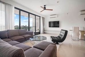 A modern living room with a sectional sofa, a glass coffee table, a black leather armchair, and a flat-screen TV mounted on the wall.