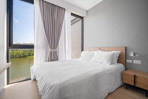 A bedroom with a large window, a bed with white linens, a wooden nightstand, and a view of greenery. - Cassia Residences, Three-Bedroom B Premium Lake (Choeng Thale)