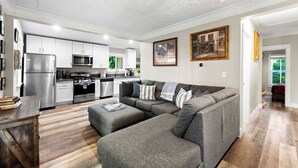 Una sala moderna con un sofá seccional, una mesa de comedor y una zona de cocina con gabinetes blancos y electrodomésticos de acero inoxidable.