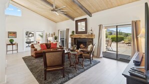 A spacious living room with a fireplace, a ceiling fan, and a large window overlooking a garden. - Club Ironwood Resort Access, Desert Garden & Golf (Carefree)