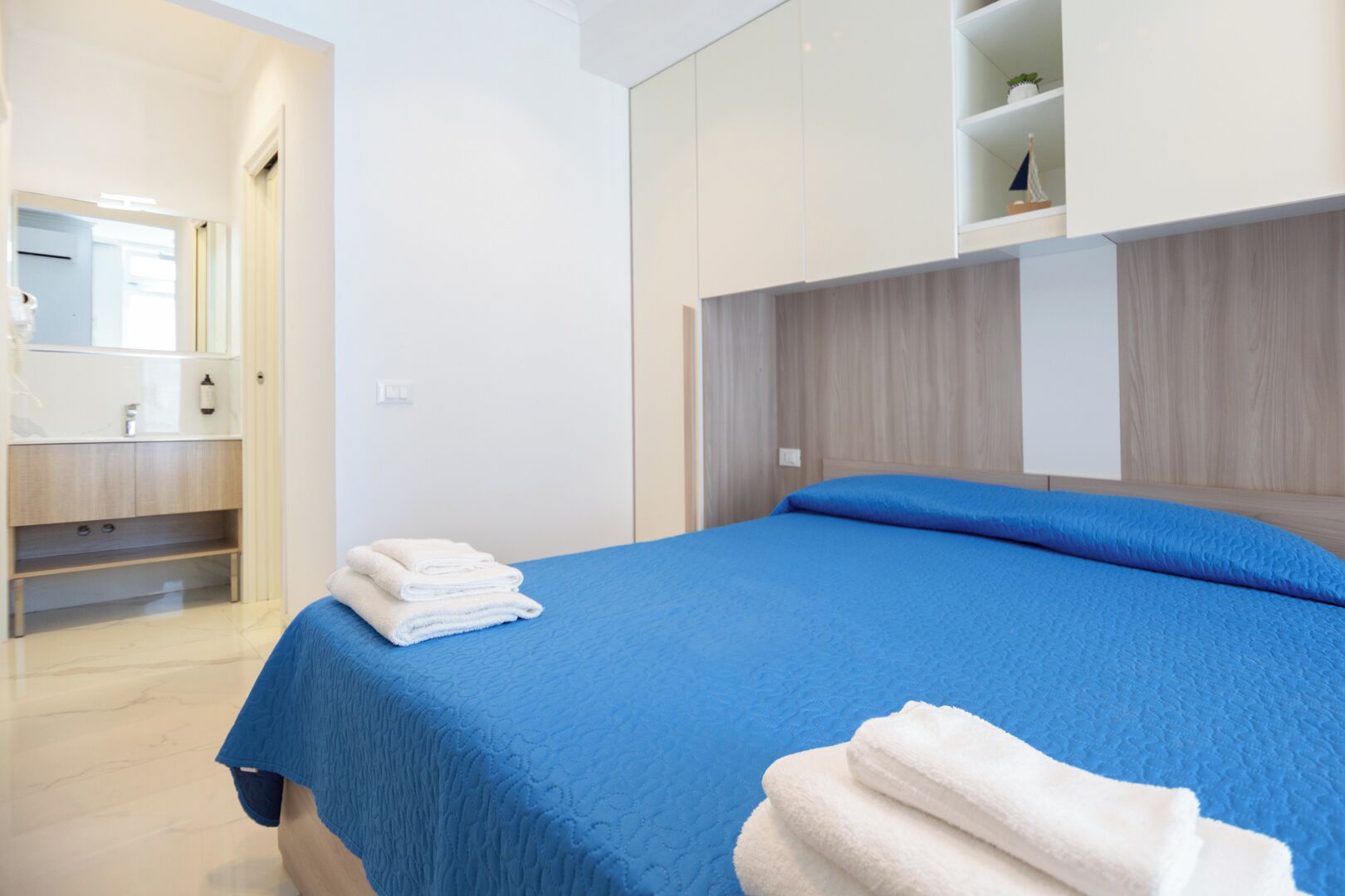 Quarto de hotel moderno com cama, penteadeira com espelho e prateleira embutida com objetos decorativos.