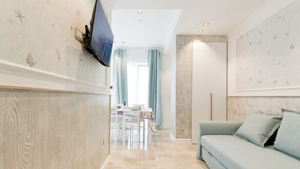 A modern living room with a sofa, a flat-screen TV mounted on the wall, and a dining area with a table and chairs.