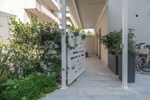 A narrow pathway flanked by greenery and a white gate, leading to a building with a balcony.