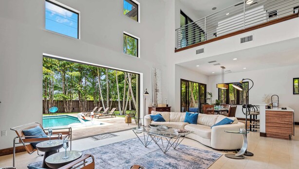 Un vaste espace de vie avec une grande fenêtre donnant sur une piscine, un coin salon confortable avec un canapé et des fauteuils, et une cuisine équipée d’une table à manger et d’un bar.