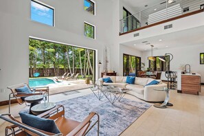 A spacious living area with a large window overlooking a pool, a cozy seating area with a sofa and chairs, and a kitchen area with a dining table and bar. - Wilton Grand Luxury: Private Oasis w/Pool (Wilton Manors)