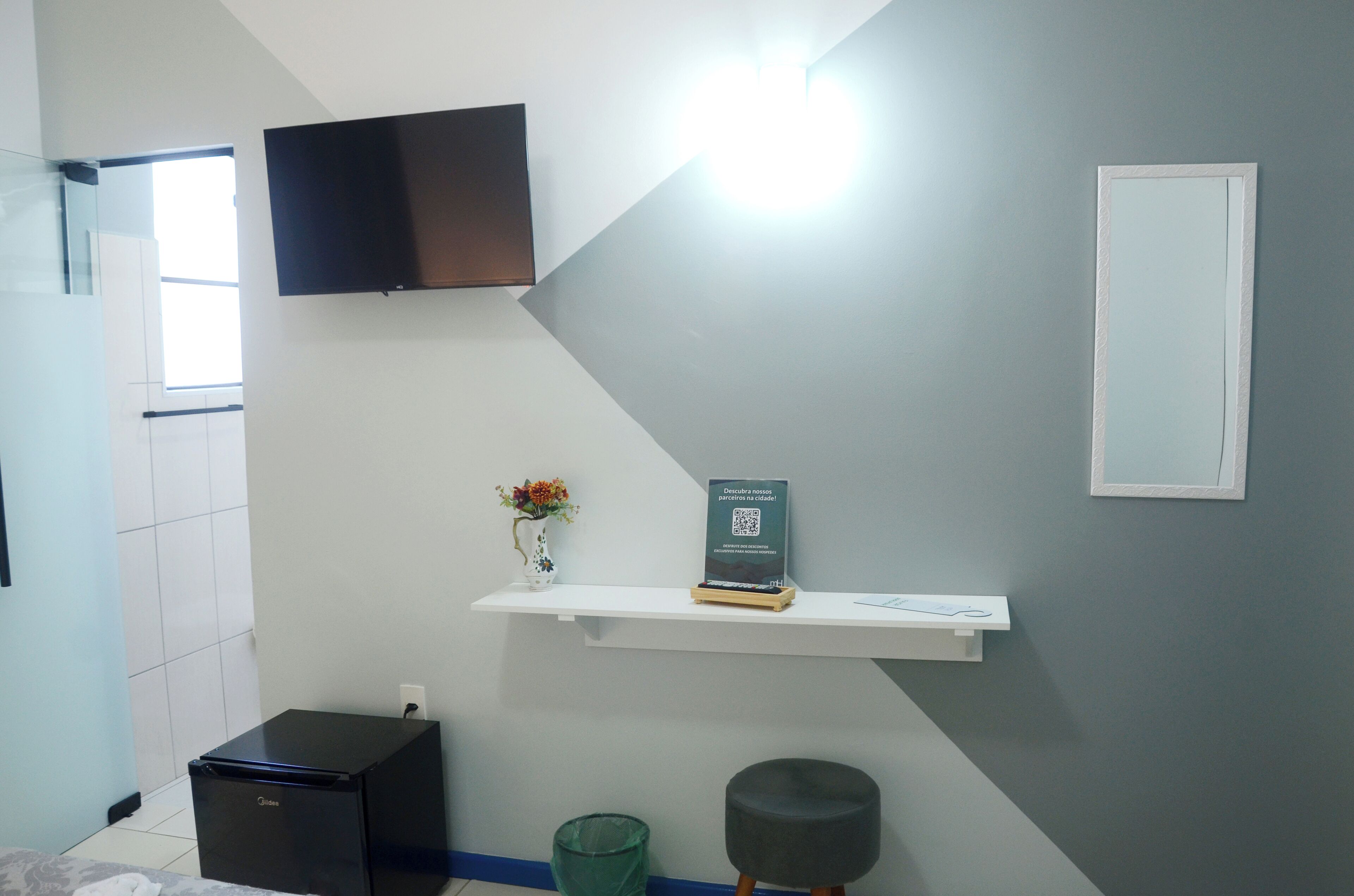 A modern room with a flat-screen TV mounted on the wall, a small black refrigerator, a white shelf with a QR code, and a mirror.
