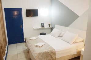 Une chambre à coucher avec un lit, une porte bleue, un téléviseur fixé au mur et une petite étagère sur laquelle se trouvent un livre et un vase.