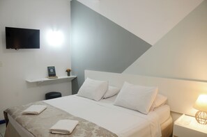 Une chambre d’hôtel moderne équipée d’un lit, d’un téléviseur à écran plat fixé au mur, d’une table de chevet avec une lampe et d’une petite étagère ornée d’un objet décoratif.