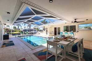 A modern indoor pool area with a glass ceiling, a dining table set for four, and a view of the pool and outdoor seating area. - Blue Skies at Bayshore: Heated Pool, Hut Tub, Dock, Bikes, Beach Gear! (Cape Coral)