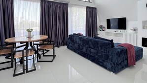 Un salon moderne avec un plafond haut, de grandes fenêtres, un canapé bleu foncé, une table à manger avec des chaises et un téléviseur à écran plat.