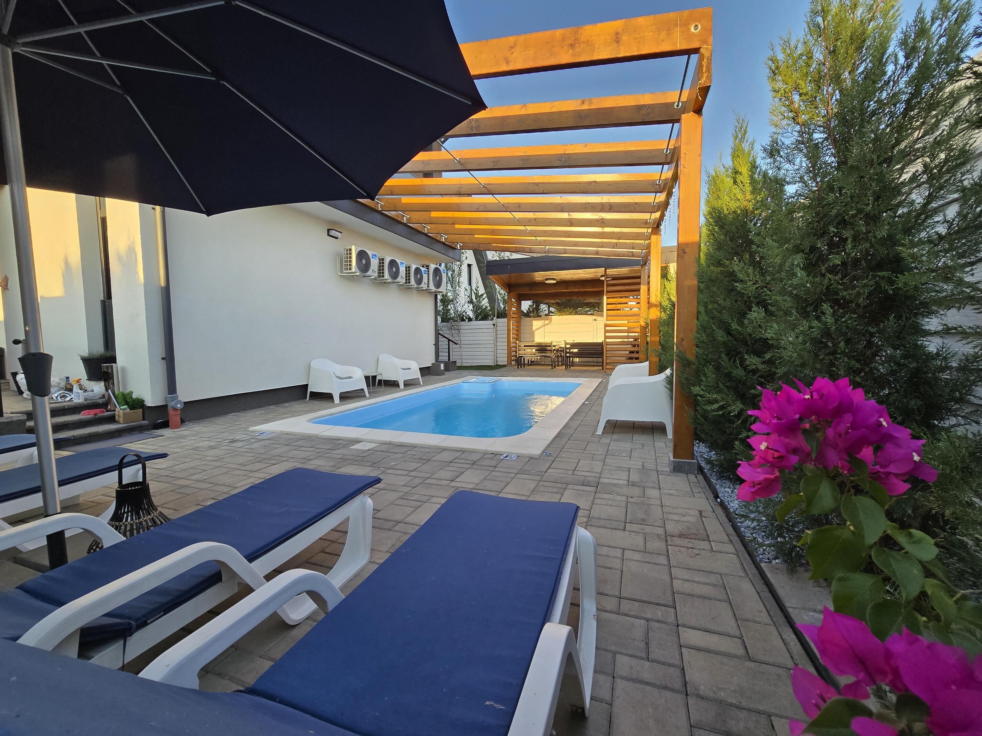 A pool area with lounge chairs and umbrellas, surrounded by a wooden pergola and potted plants.
